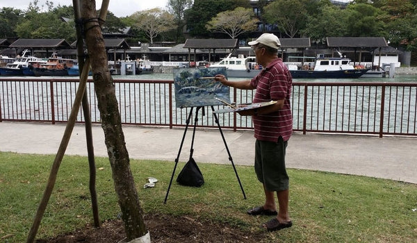 Our artist Low Hai Hong is a Singapore artist. ARTualize is the only art gallery in Singapore that represents him and his works.  A outdoor photo of Low Hai Hong painting an oil painting mounted on a small easel under a tree painting boats on a river.