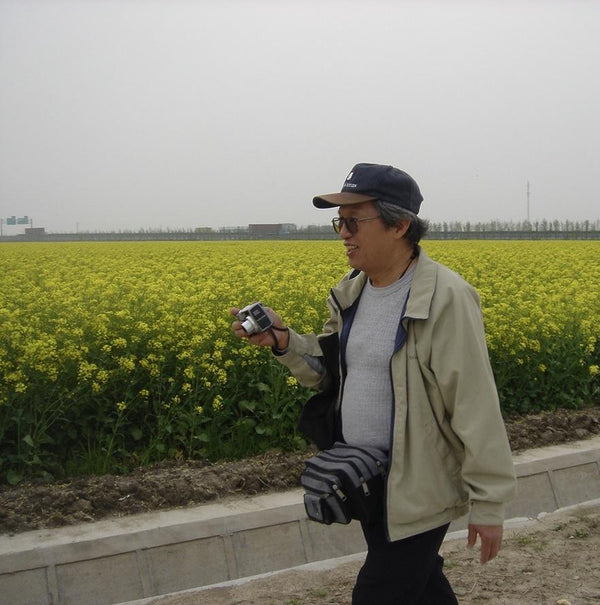 Our artist Low Hai Hong is a Singapore artist. ARTualize is the only art gallery in Singapore that represents him and his works.  Low Hai Hong is walking with a huge smile, holding a camera in his hand, walking past a huge field full of yellow flowers.